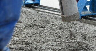 Nahaufnahme von frisch eingebrachtem Beton, der aus einem Rohr auf eine Baustelle gegossen wird. Im Vordergrund ist ein Teil einer blauen Arbeitskleidung zu sehen, im Hintergrund Baugeräte. Die Konsistenz des Betons ist grobkörnig und feucht.

                                    